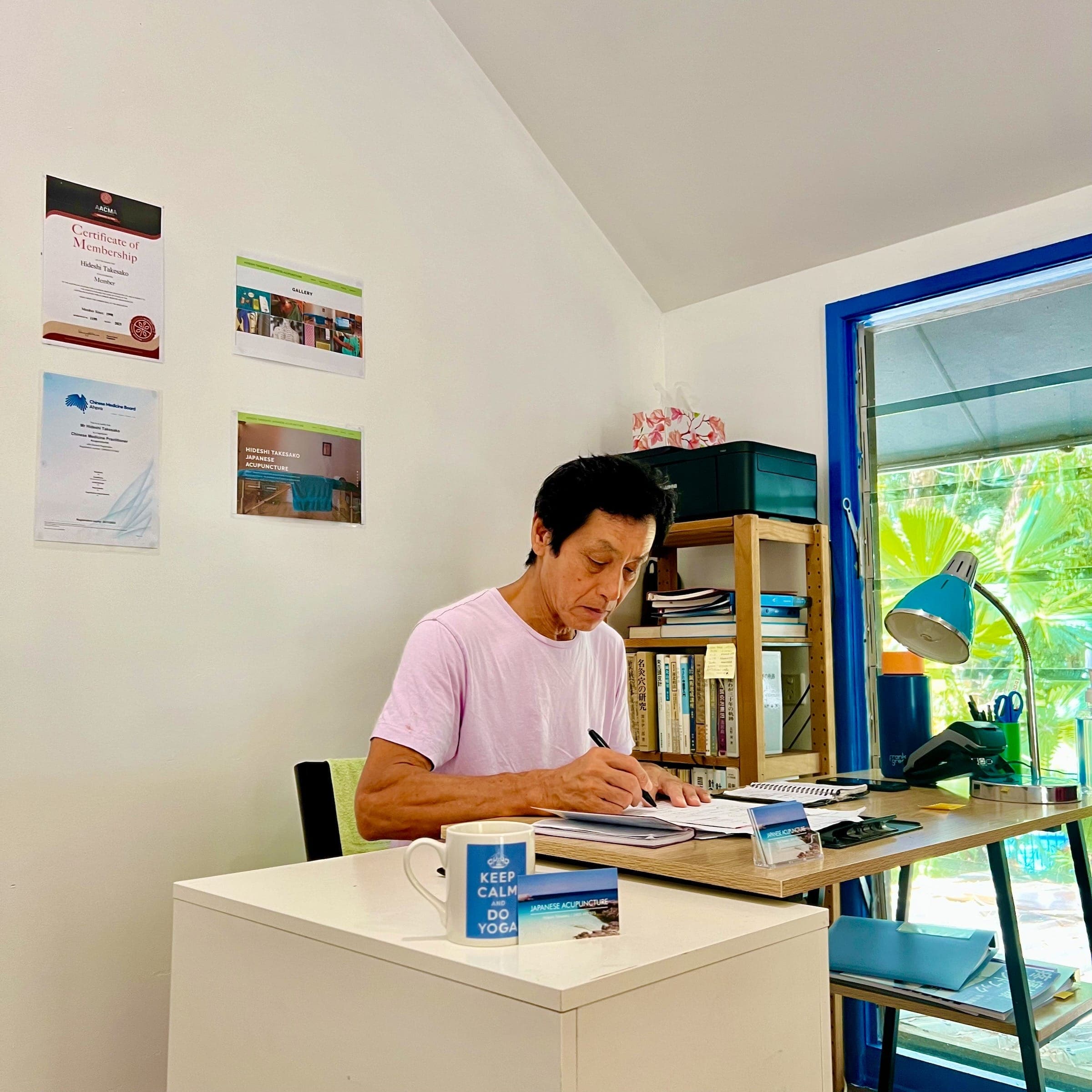 Hideshi reviewing patient notes at his desk, with professional certificates visible in the background